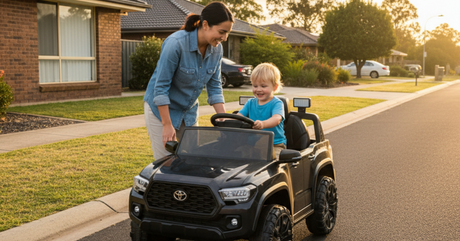 A smiling woman helping a young boy who is sitting in a black Toyota Tacoma ride-on toy truck. They are on a suburban street or driveway at sunset.