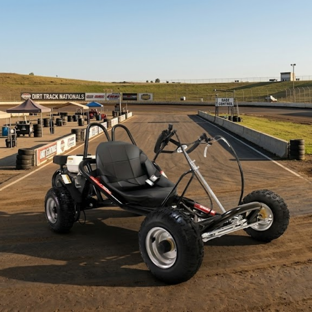 A black go-kart with a roll bar and large off-road tires parked on a dirt track at an outdoor racing facility under a clear blue sky.