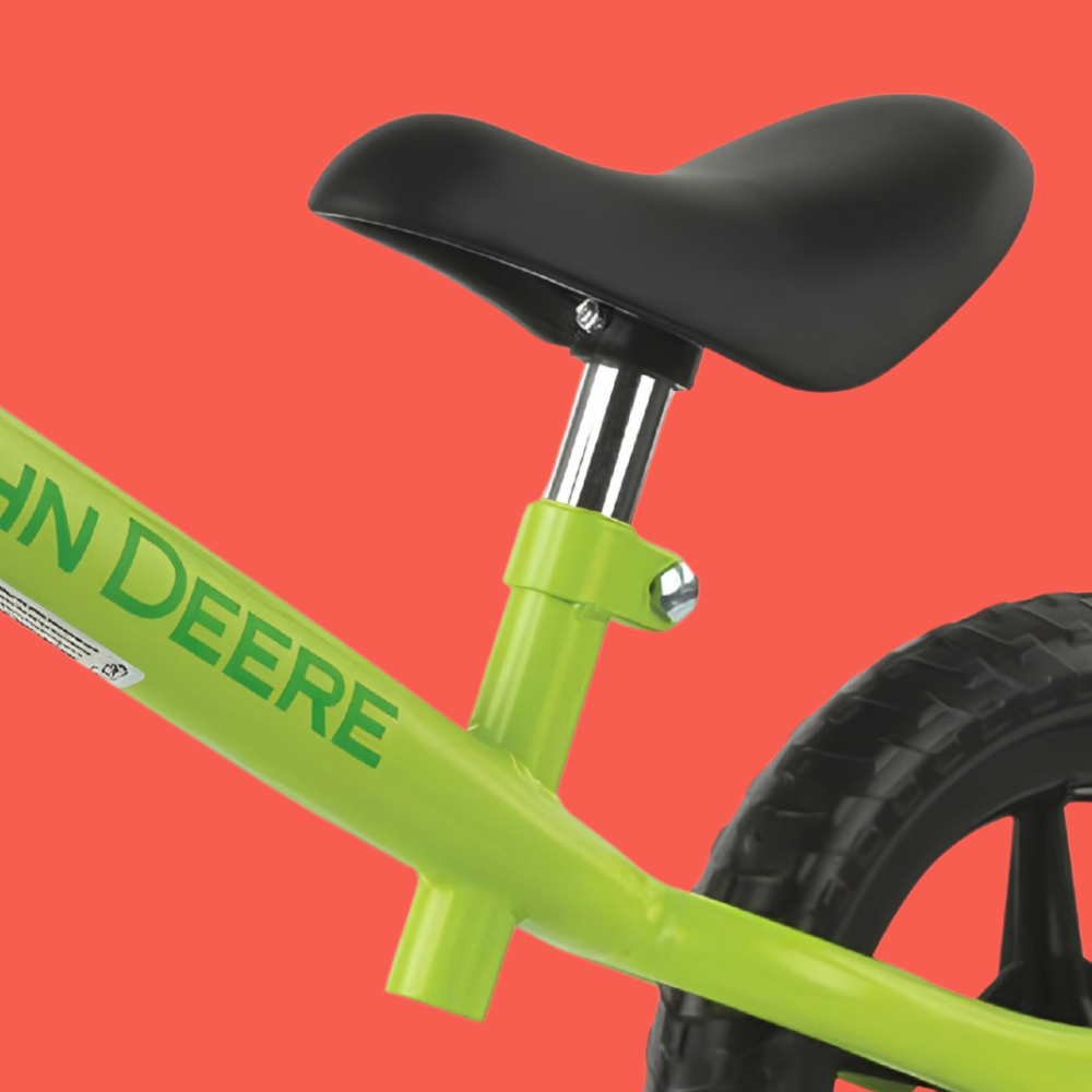 Close-up of a bright lime green balance bike frame with a black saddle and black tire, shown against a solid coral-red background. The brand name 'John Deere' is printed in green on the frame.