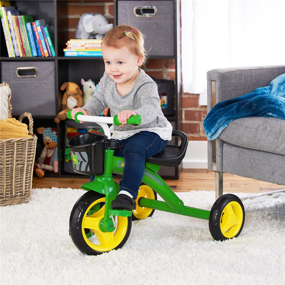 Smiling toddler riding a John Deere steel tricycle with green frame and yellow wheels inside a living room.