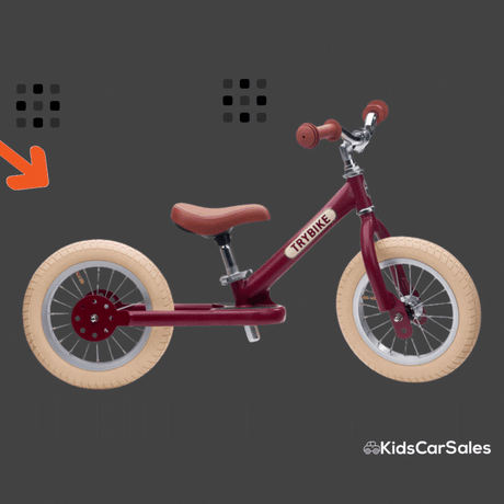 A maroon Trybike balance bike with cream tyres and brown seat and grips is shown on a dark grey background with a small orange arrow pointing to the rear wheel. The Kids Car Sales logo appears in the bottom-right corner