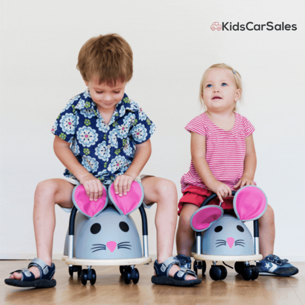 Two small children sit side-by-side on Wheely Bug Mouse ride-on toys with large pink ears, indoors on a wooden floor.