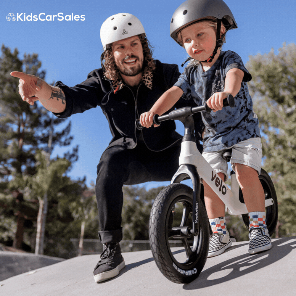 A man in a white helmet points as a child rides a white Yvelo balance bike down a concrete ramp. Both wear helmets.
