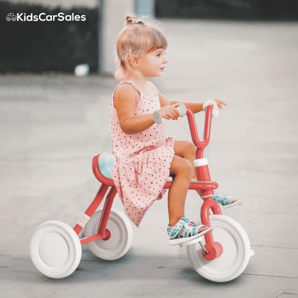 A young girl in a pink polka dot dress pedals a red and tan Yvolution Velo Trike across a smooth, grey outdoor pavement.