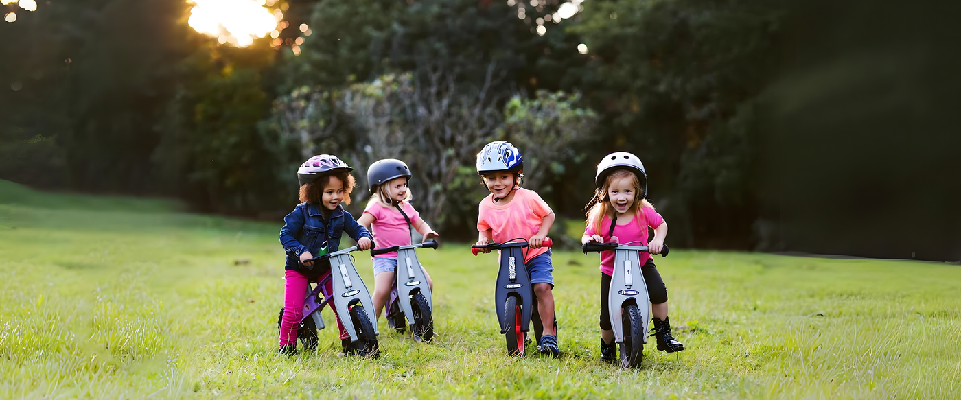 Four children wearing helmets are riding balance bikes outdoors on a grassy field, smiling and enjoying their time together