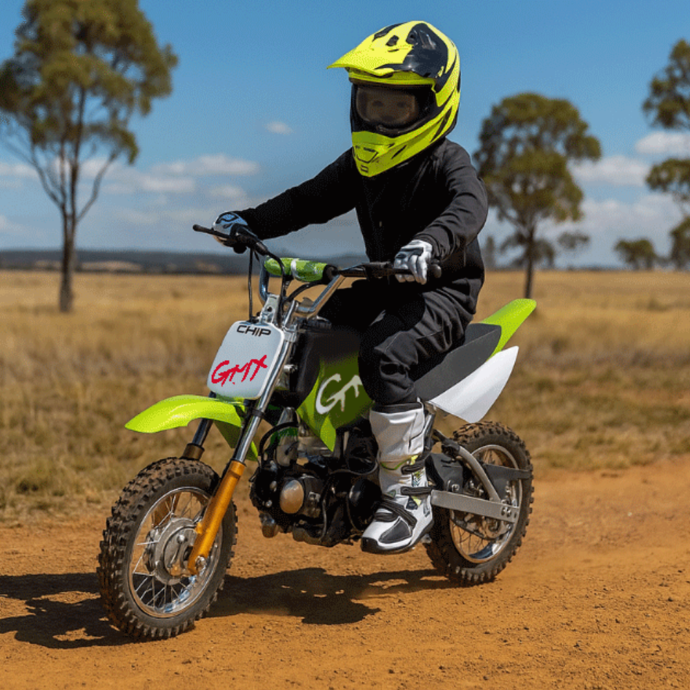 Action shot of a child riding the green GMX dirt bike on a dry dirt trail, wearing a yellow helmet and black riding gear.