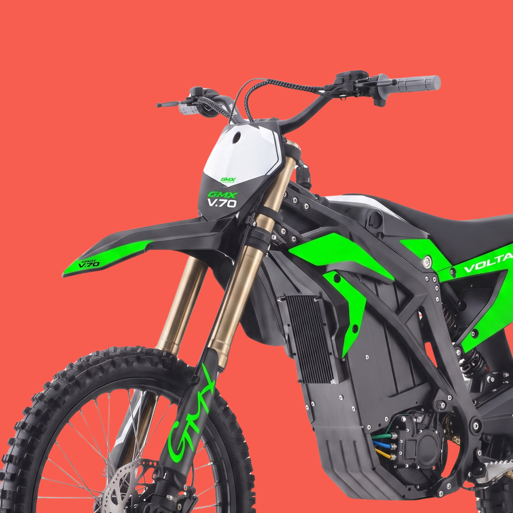A close-up studio shot of a Volta V70 electric dirt bike against a solid red-orange background. The bike features a black frame, a white front number plate, gold front forks, and vibrant green accents on the body and fender.