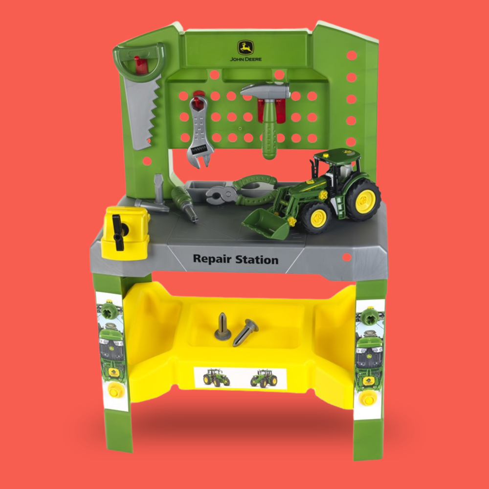 John Deere toy repair station playset with a green and gray workbench, yellow tools, hanging wrenches, a toy tractor, and small hardware pieces, shown on a solid coral-red background.