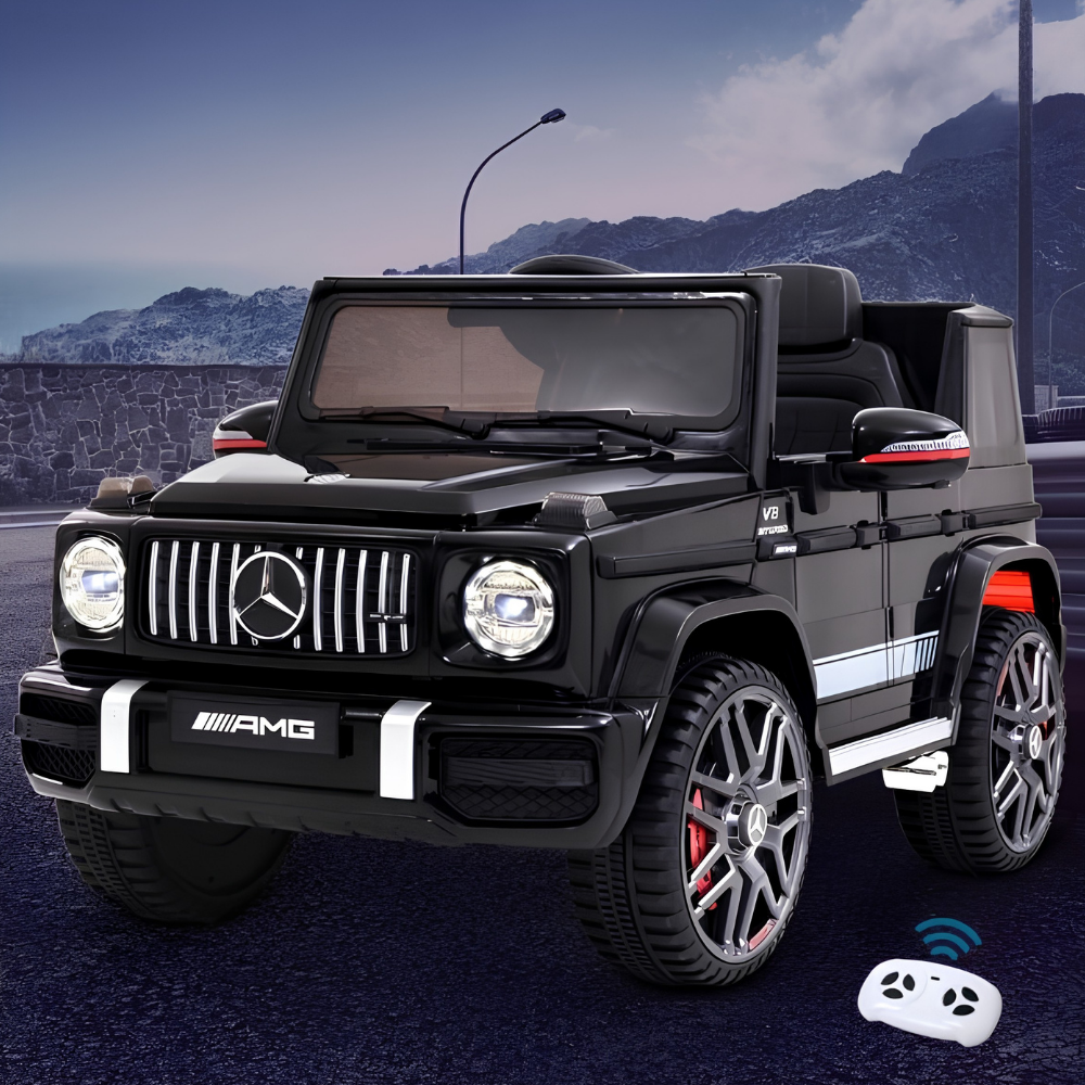 A black Mercedes-Benz G-Class style children's ride-on vehicle with chrome grille, round headlights, and large wheels, shown outdoors on a road with mountains in the background. A small white remote control is visible in the lower right corner.