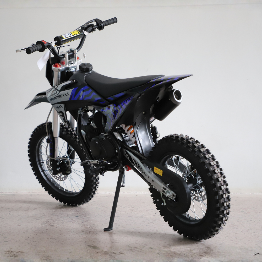 A black and blue off-road dirt bike with knobby tires, a black seat, and a visible exhaust pipe, photographed indoors against a plain light gray wall.
