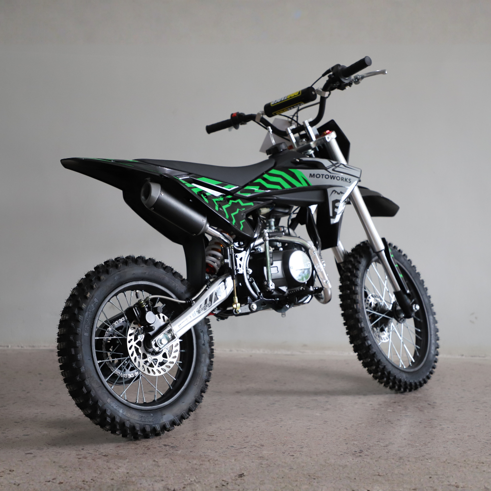 A black and green mini dirt bike with off-road tires, a black exhaust, and silver forks, photographed in a studio setting against a neutral gray background.
