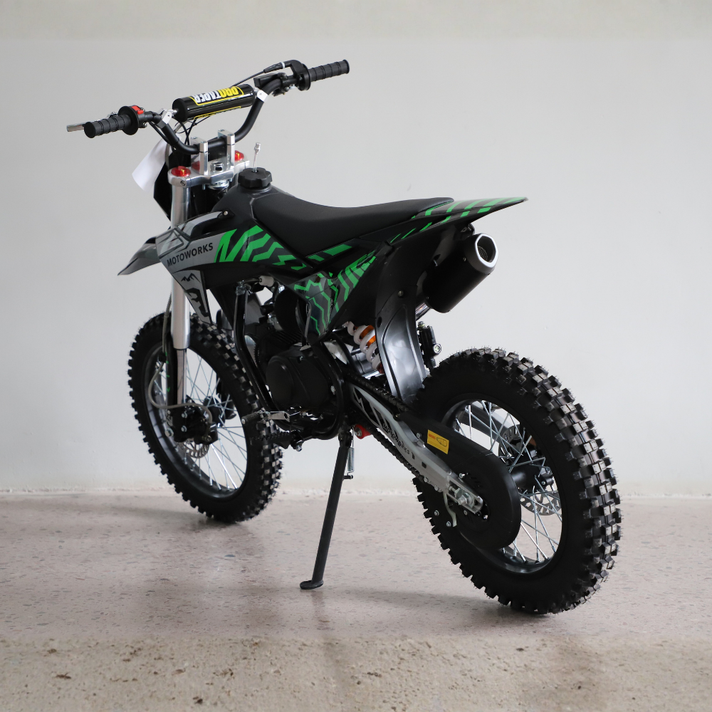 A youth dirt bike with a black seat and bodywork featuring green graphic patterns, standing on its kickstand in a studio setting.