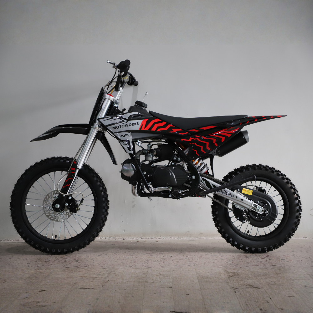 A small off-road motorcycle with a black and red patterned seat, black fenders, and a silver engine, photographed in profile against a plain light gray wall.