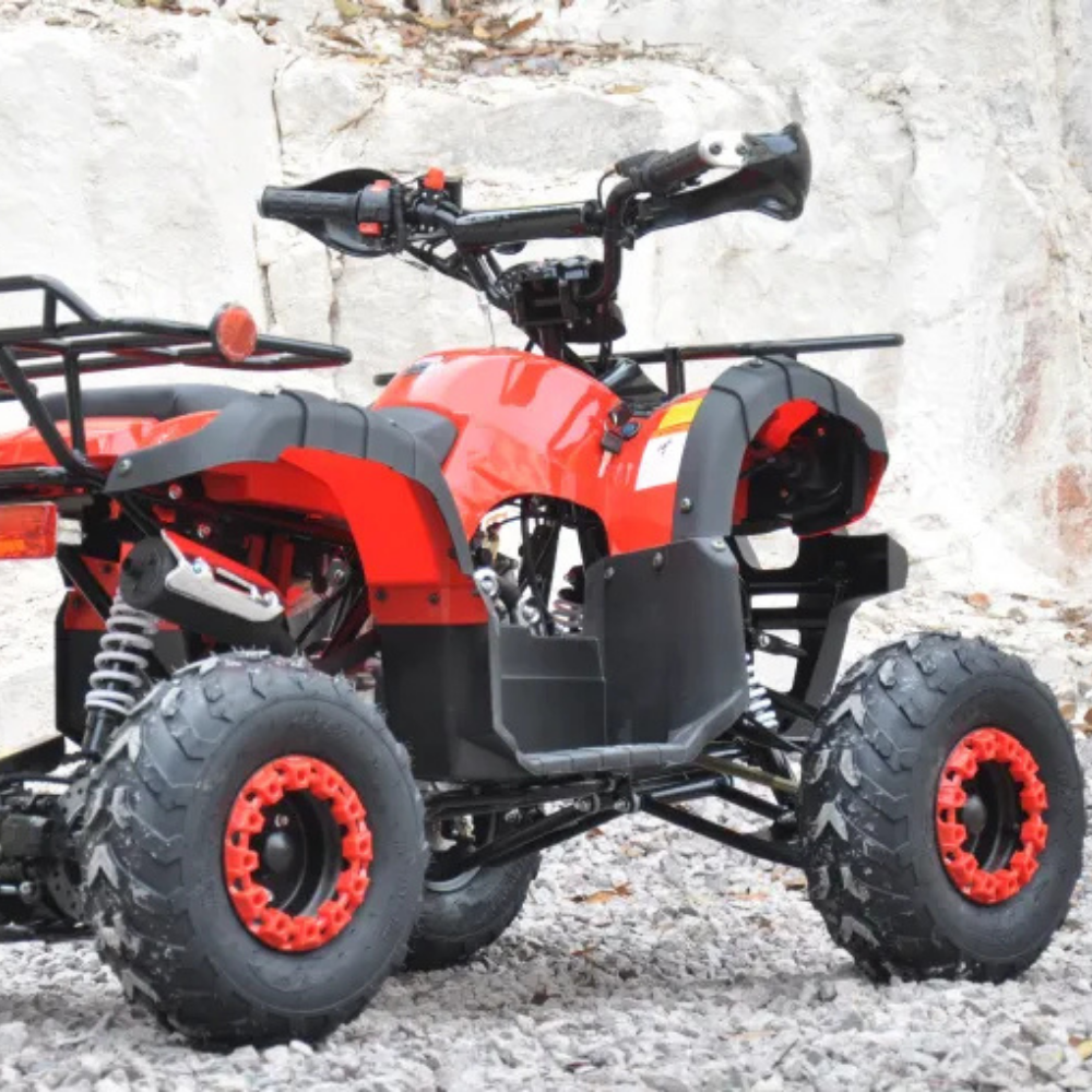 Red and black kids quad bike on rocky ground, front-left angle with visible suspension and tyres.