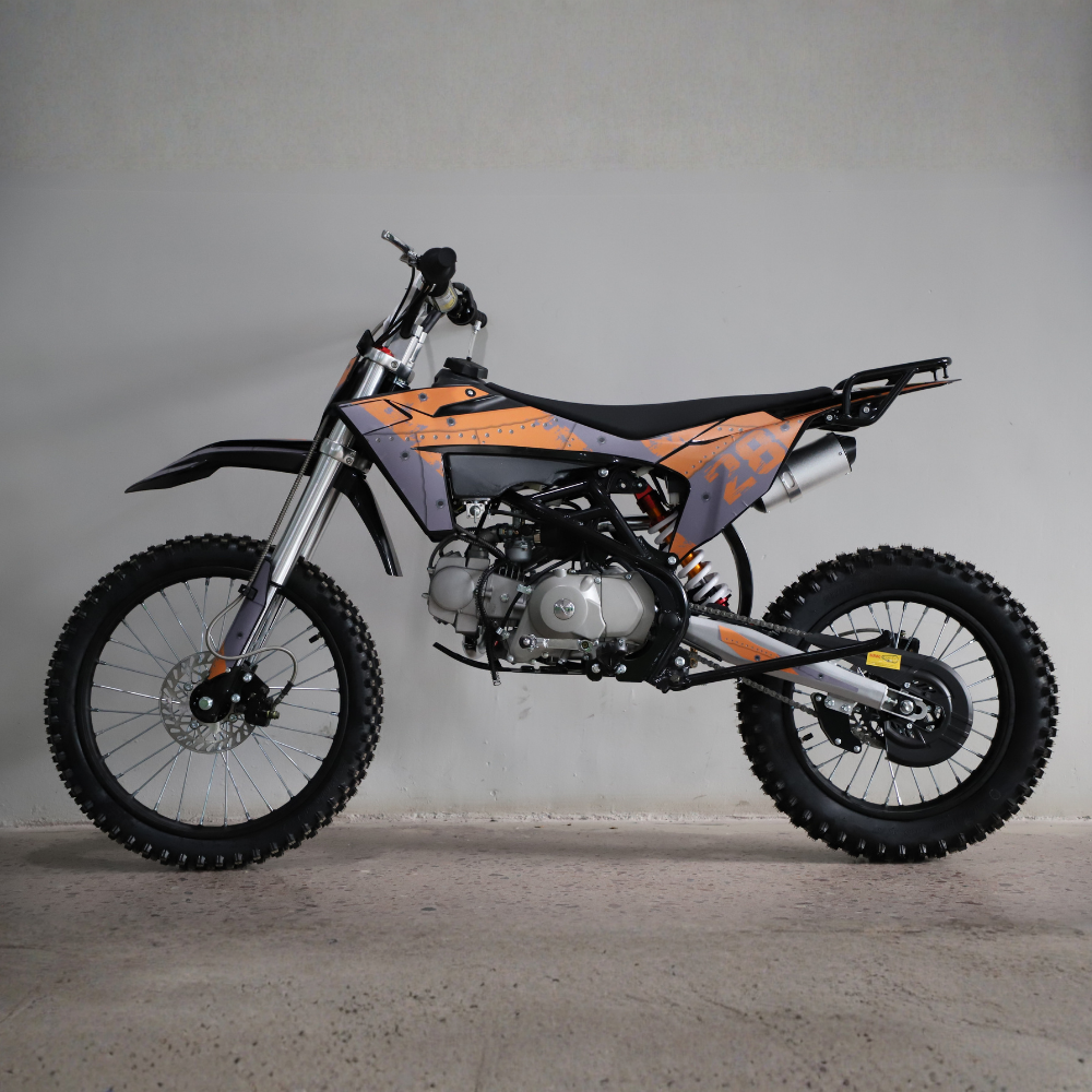 A dirt bike with orange and gray graphics, black seat, and silver engine components, photographed in profile against a plain gray wall and concrete floor.