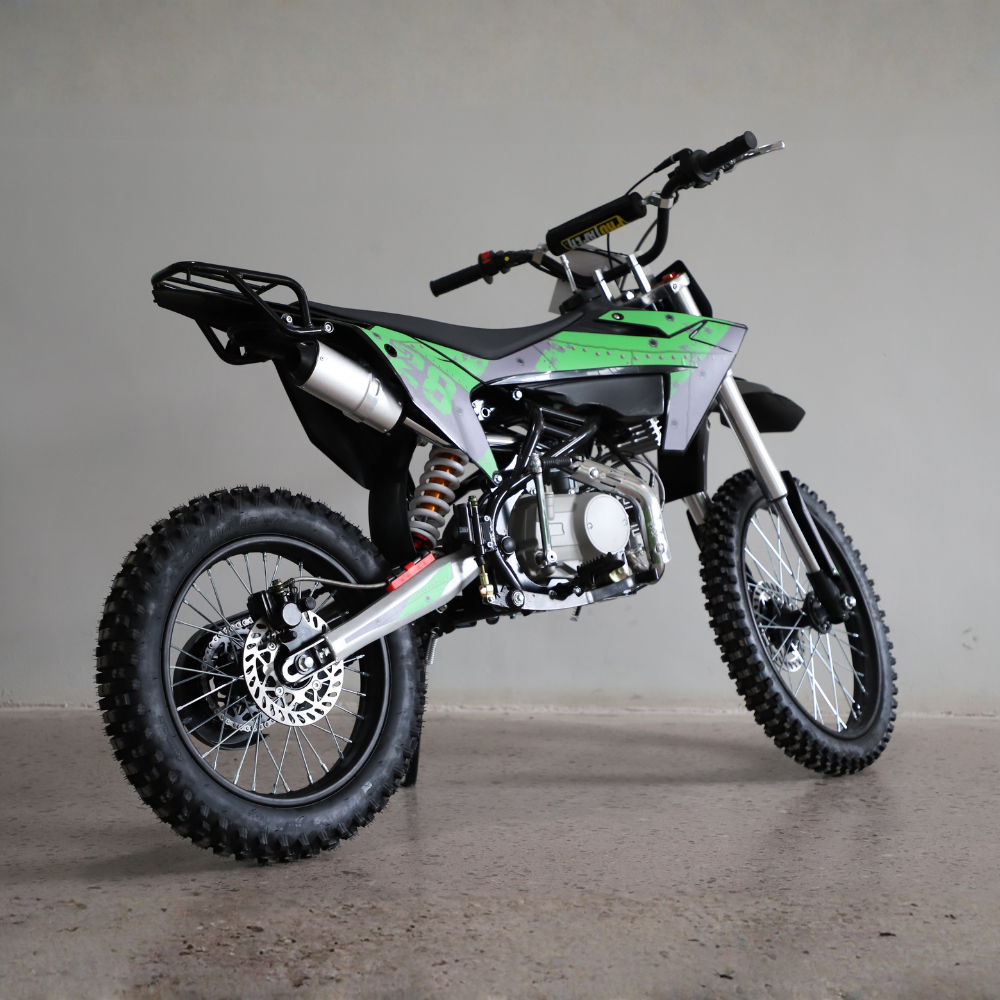A green and black mini dirt bike with a silver exhaust and black knobby tires, photographed on a concrete floor against a plain gray wall.