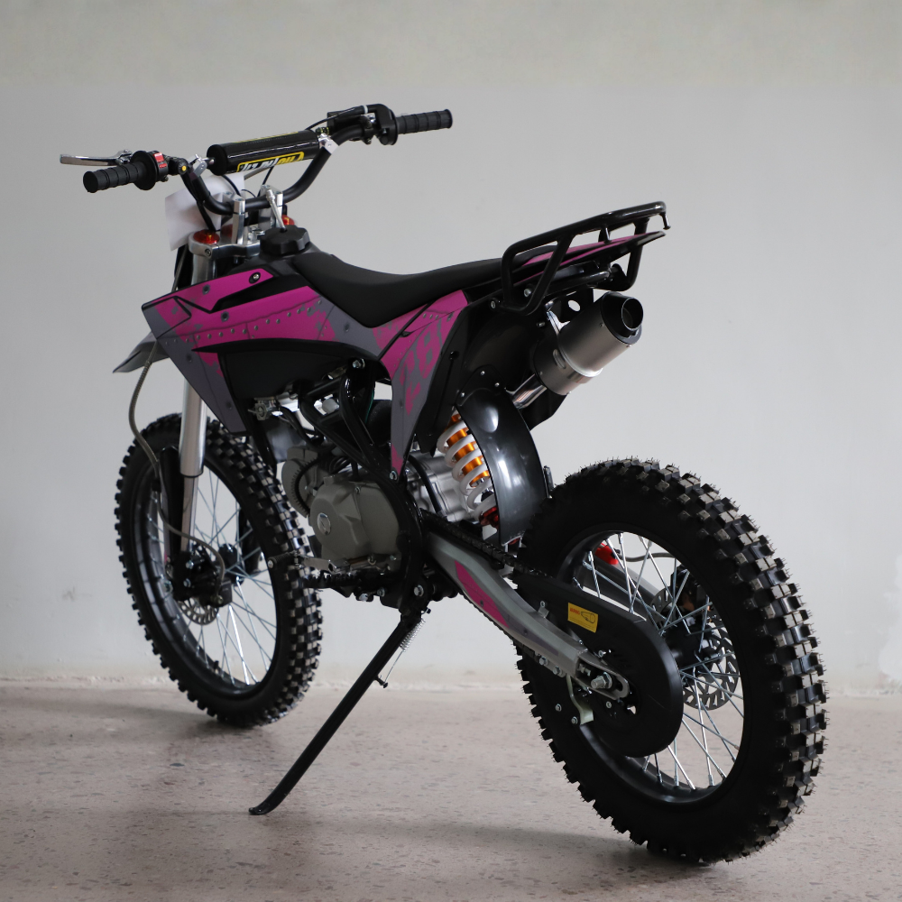 A pink and gray dirt bike with knobby off-road tires, a black seat, and a rear luggage rack, photographed on a concrete floor against a plain light gray wall.