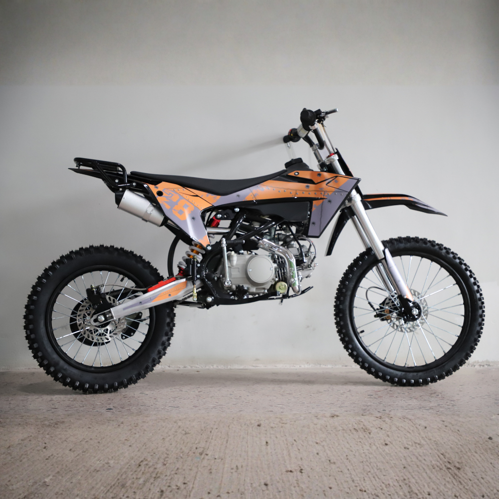 A 125cc dirt bike with orange and black graphics, silver forks, and knobby tires, photographed from a side profile against a plain concrete wall.
