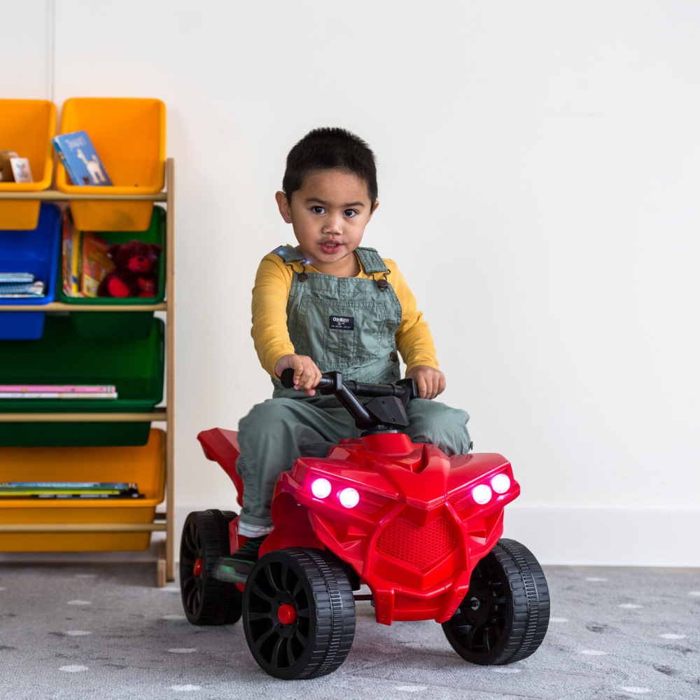 Child sitting on a red toy ATV in a room with colorful shelves.