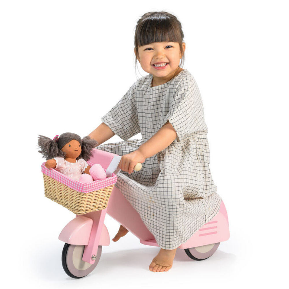 Child on a pink scooter with a basket containing a doll and toys, wearing a checkered dress.