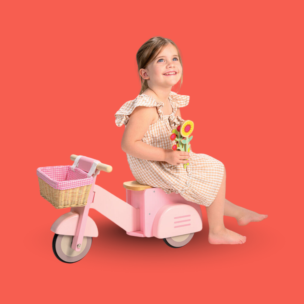 Young girl sitting on a pink toy scooter with a basket against a red background