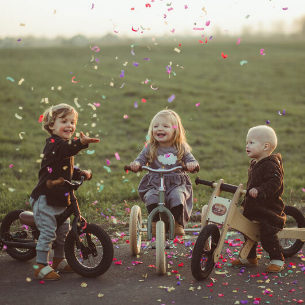 Three children on balance bikes with confetti in a field