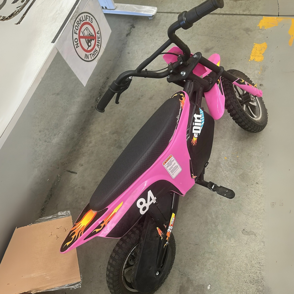 Angled view of pink electric dirt bike parked indoors, showing worn seat and colourful decals on body.