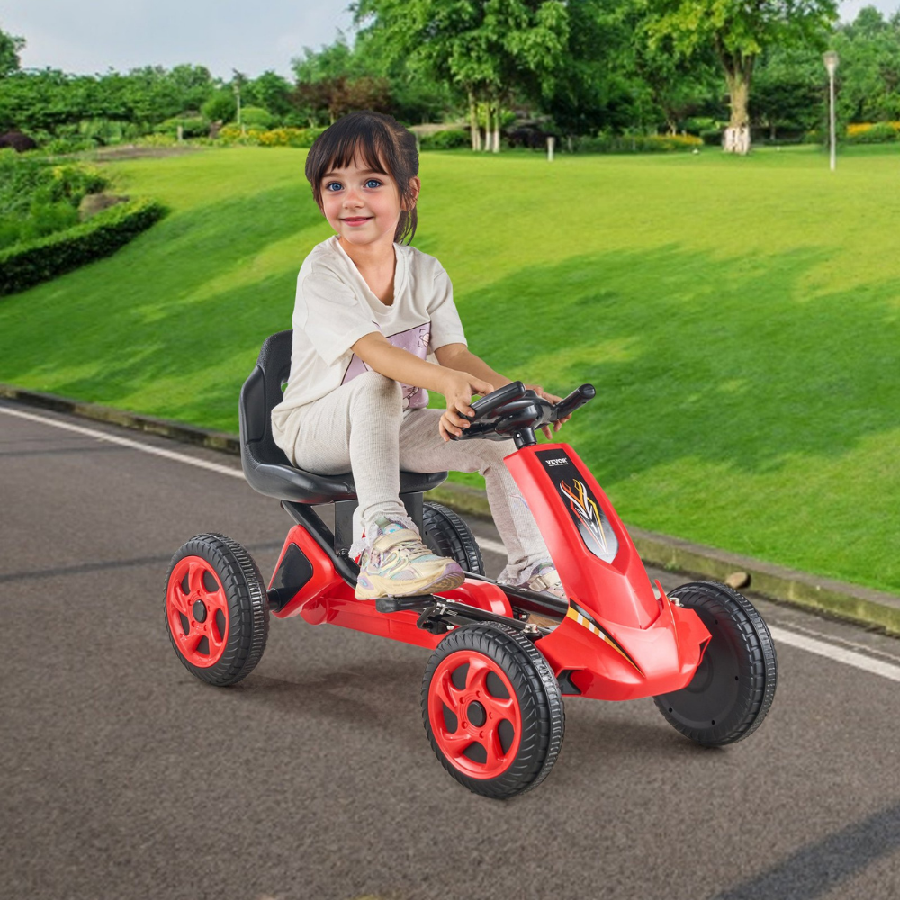 Child riding a red pedal go-kart on a paved path with green grass and trees in the background.