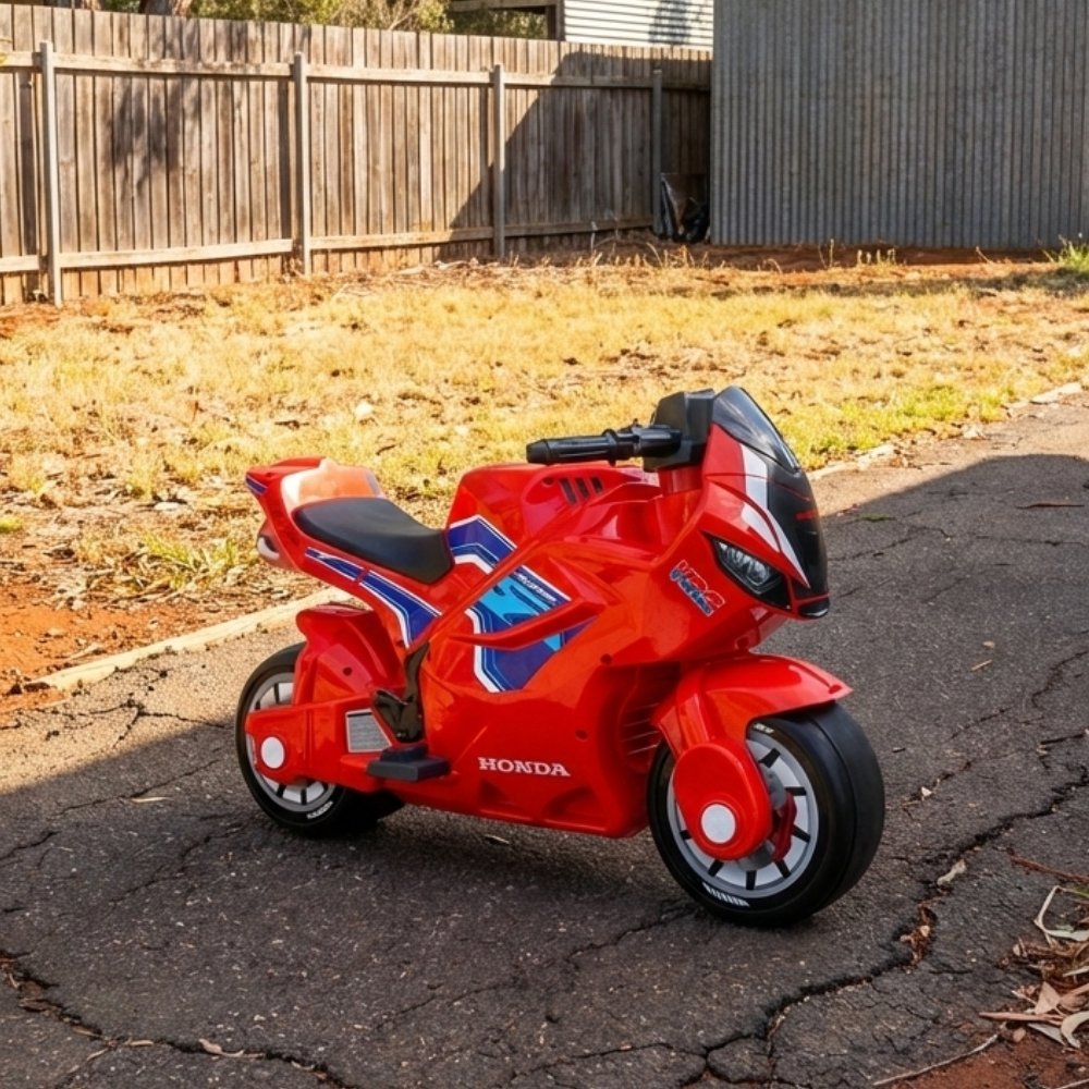 Red Honda toy motorcycle on a cracked pavement with a wooden fence and grass in the background.