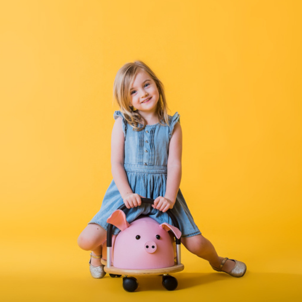 Young girl in a denim dress sitting on a pink pig-shaped suitcase against a yellow background