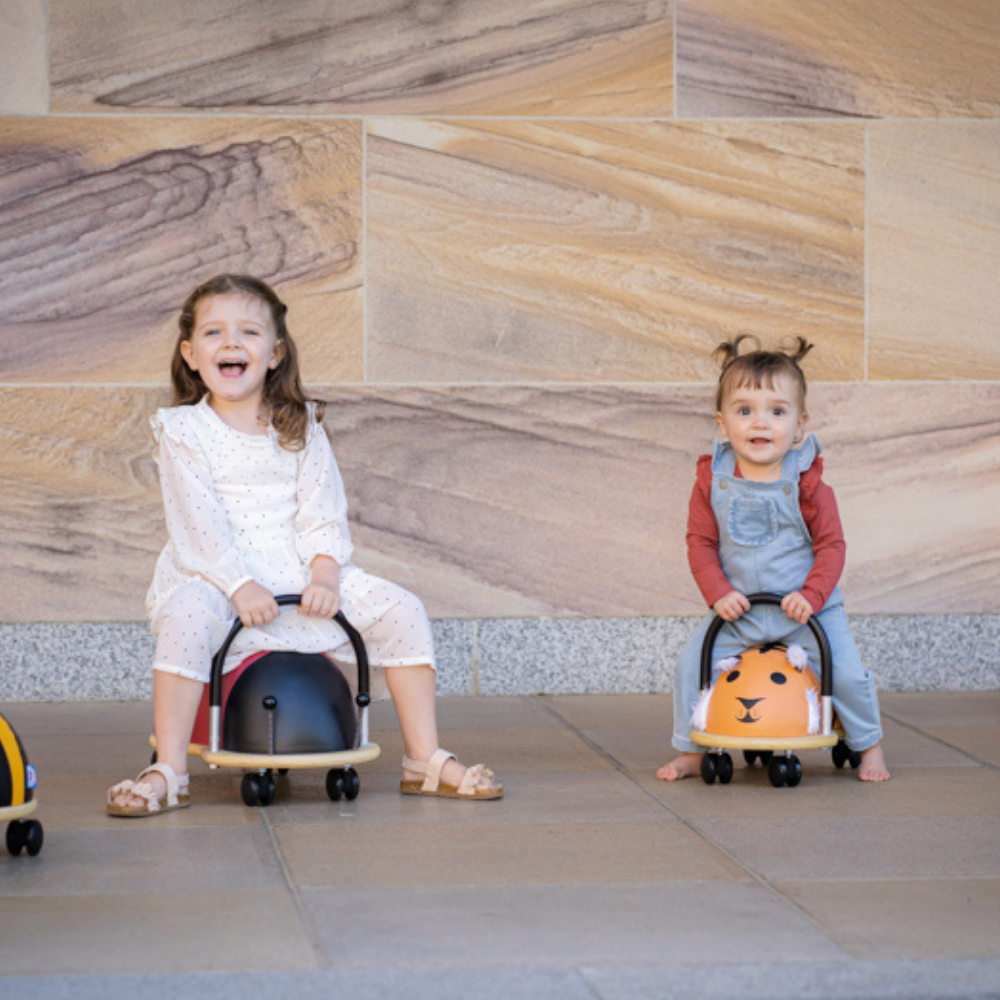 Two children sitting on small, round, black and orange ride-on toys against a stone wall.