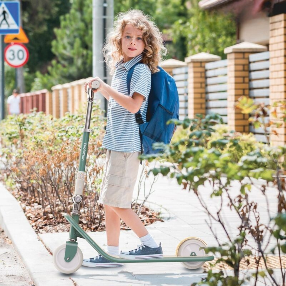 Child with a scooter and backpack standing on a sidewalk with greenery and a building in the background.