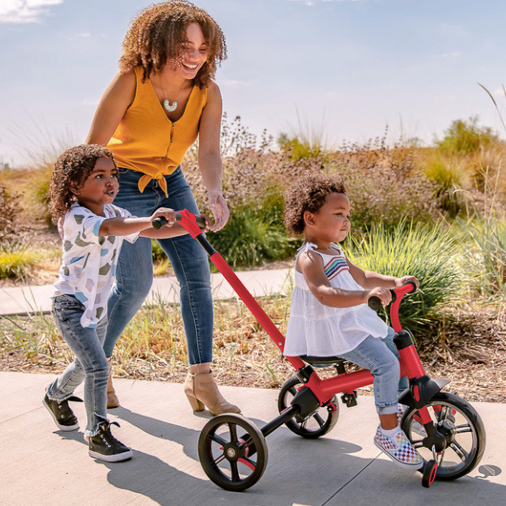 Woman pushing a child on a red bike with training wheels outdoors.