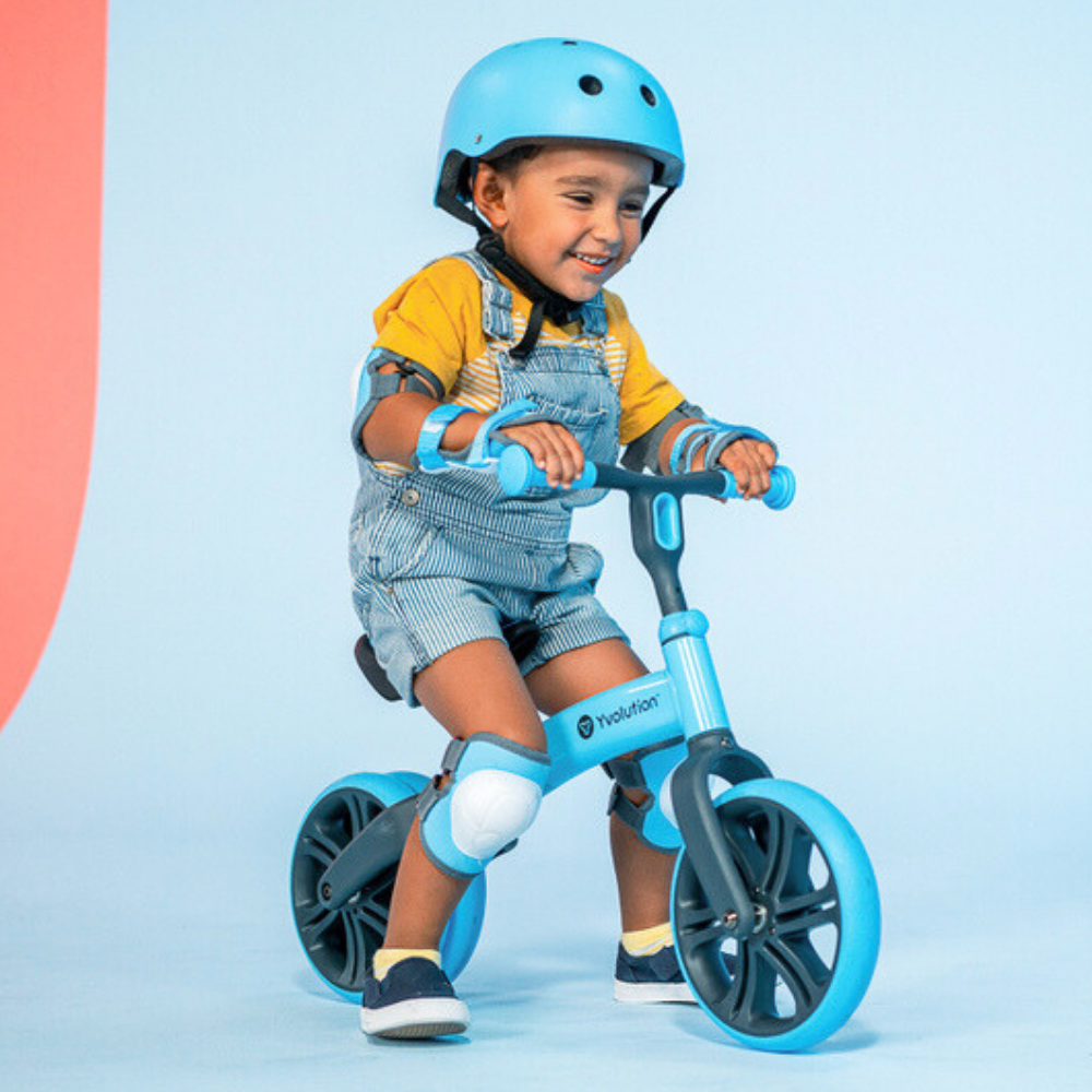 Child wearing a blue helmet and knee pads, riding a blue balance bike against a light blue background.
