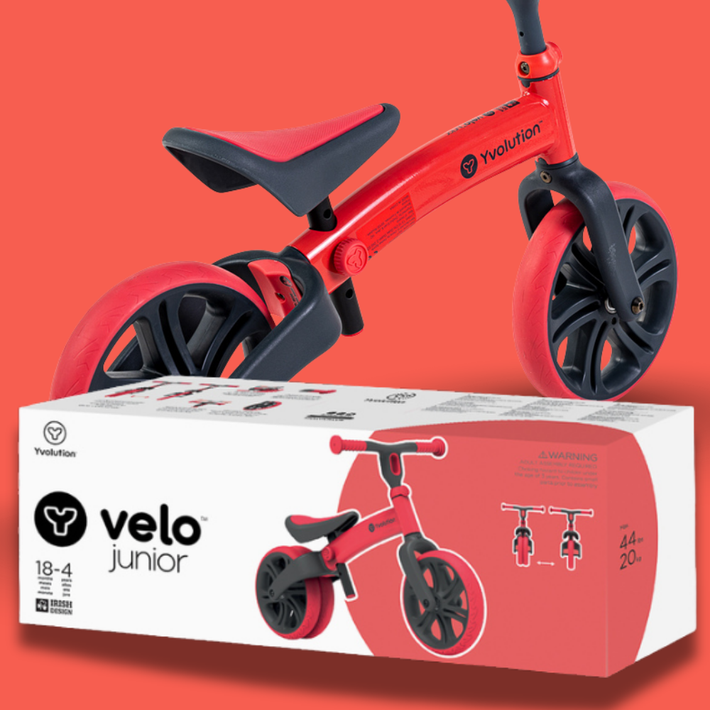 Red balance bike with black seat and wheels on a white box featuring 'velo junior' branding against an orange background.