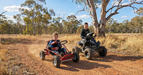 Two boys ride a red go-kart and a black ATV on a dirt track surrounded by trees on a sunny day.