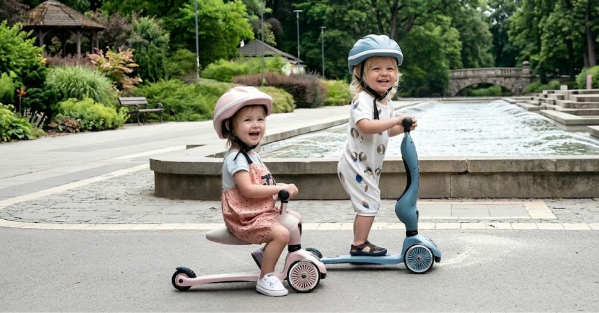 Two toddlers wearing helmets smile while riding a pink ride-on toy and a blue scooter next to a park fountain.