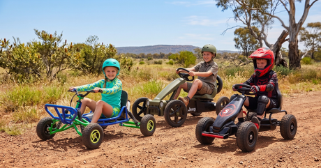 Three kids enjoy a sunny day outdoors, each riding a different style of colorful go-kart on a dusty dirt trail.