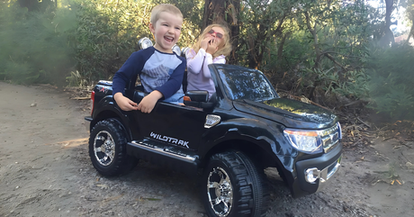 Two children laughing while riding together in a black two-seater Wildtrak ride-on ute for kids.