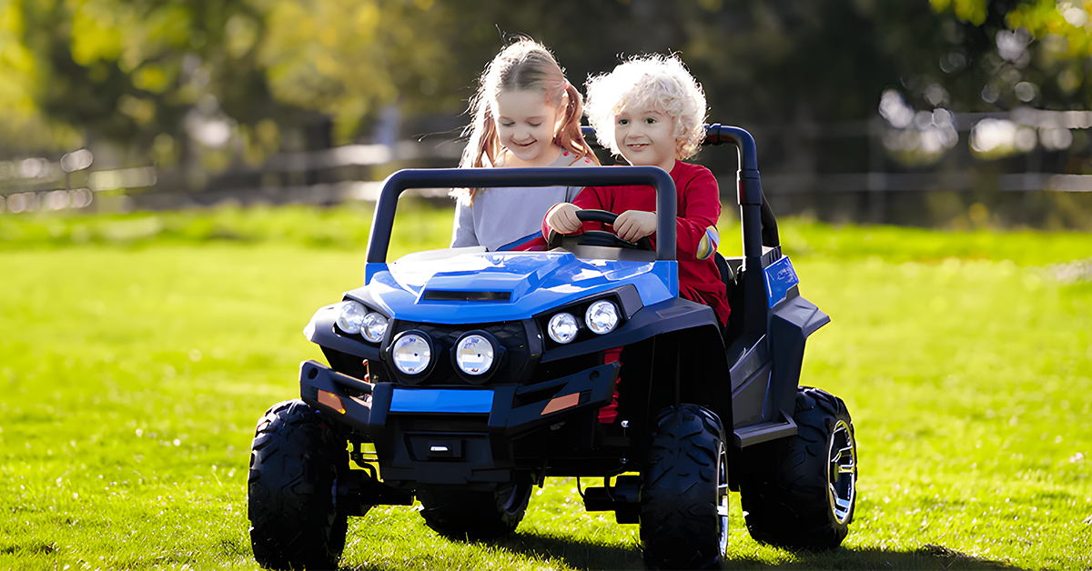 Two children are enjoying a ride in a blue electric toy car on a grassy field. The child in the driver's seat is steering the vehicle, while the other is sitting next to them