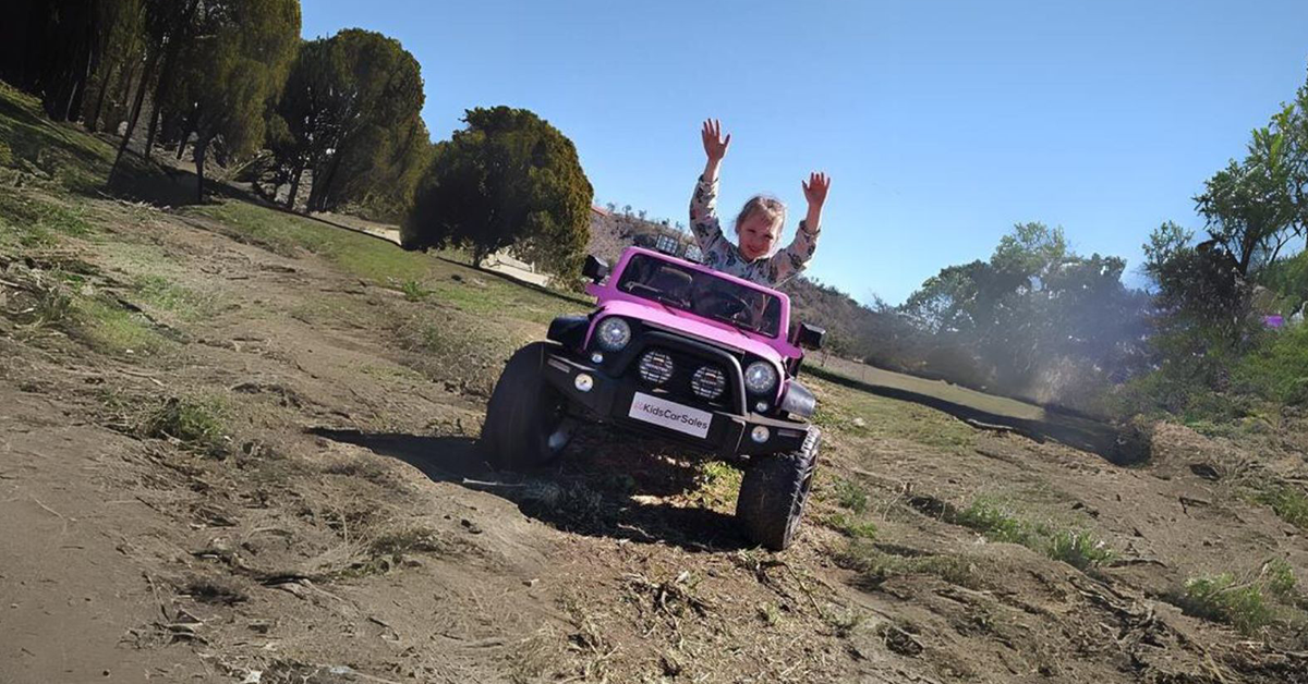 A child driving a pink toy jeep off-road, arms raised in excitement, with trees and a dirt path in the background.