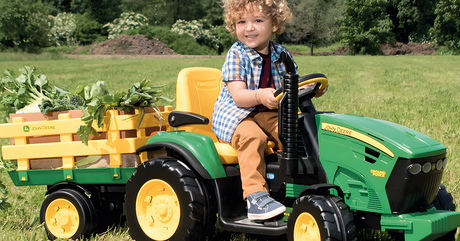 A child riding a green and yellow John Deere toy tractor with a trailer filled with plants in a field.