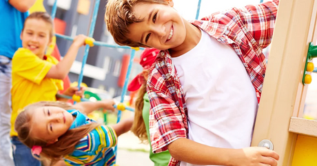 A boy in the foreground, wearing a red checkered shirt over a white T-shirt, is smiling widely while holding onto playground equipment or a structure