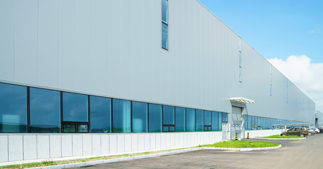 A large, modern warehouse facility with reflective glass windows, situated on a spacious lot under a bright blue sky.