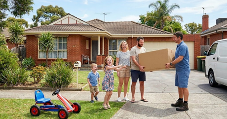 A delivery driver hands a large cardboard box to a smiling family outside their brick suburban house on a sunny day.