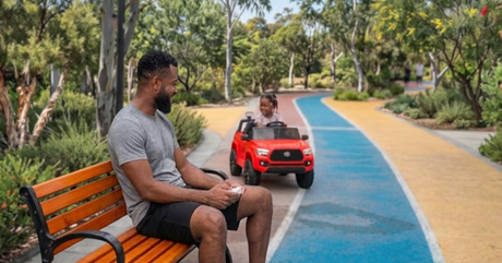 A man sits on a bench holding a remote controller, watching a toddler drive a red ride-on toy truck down a park pathway.