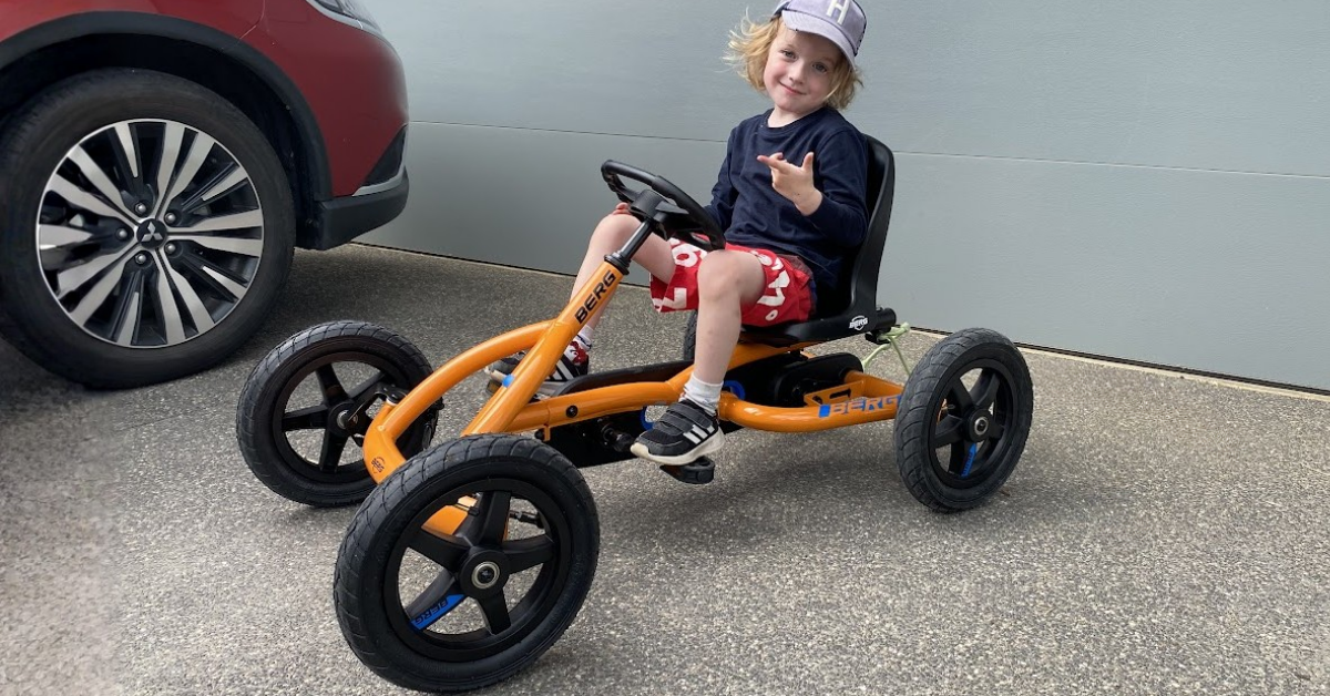 A blonde child in a hat and red shorts sits on an orange BERG pedal go-kart on a driveway near a car.