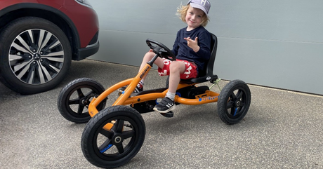 A blonde child in a hat and red shorts sits on an orange BERG pedal go-kart on a driveway near a car.