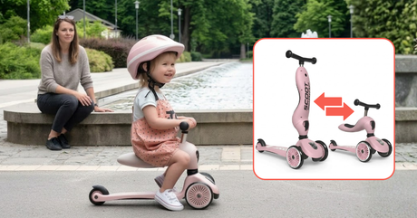 A smiling child wearing a pink helmet rides a pink three-wheel seated scooter along a paved path, while an adult watches from beside a fountain.