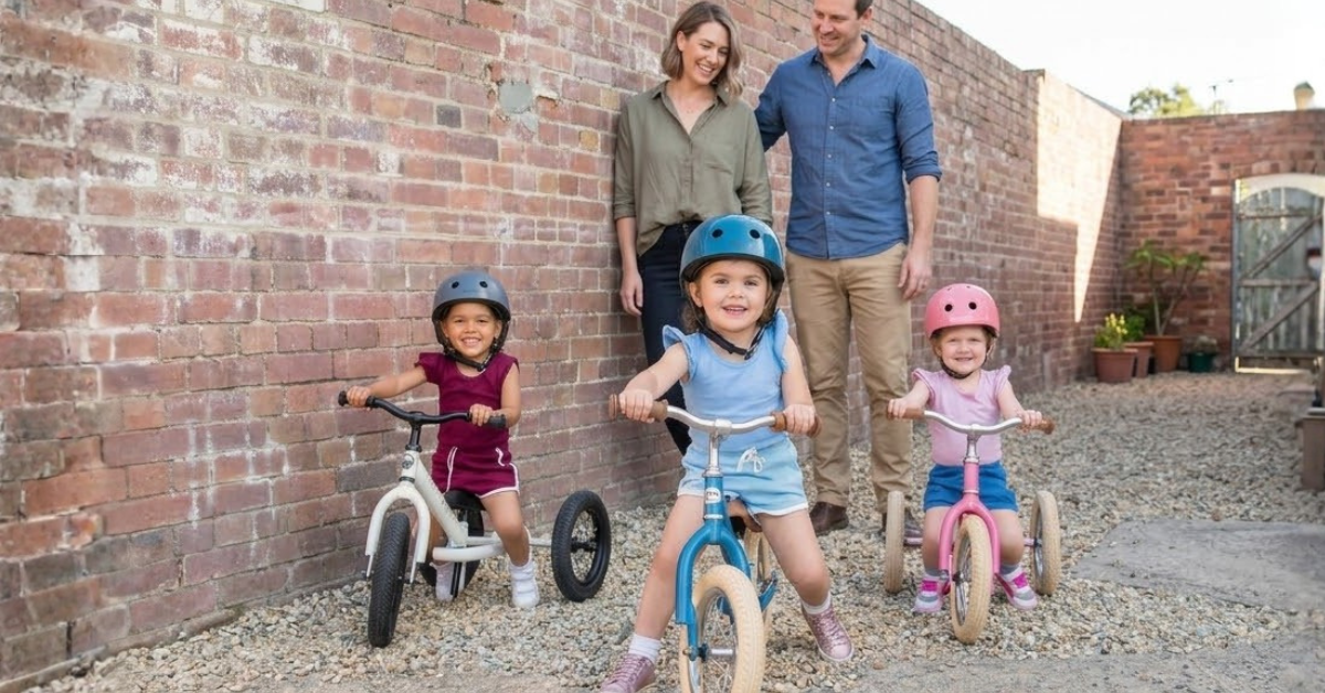 Three young kids wearing helmets ride balance bikes and trikes on gravel, with two smiling parents watching them.